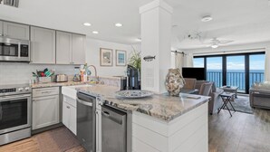 A modern kitchen with stainless steel appliances, a marble countertop, and a large island. There is a dining area with a view of the ocean. - 1201 Fall Beachfront Escape—3BR/3BA, Wraparound Balcony, Beach Service (Panama City Beach)