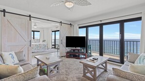 A spacious living area with a large TV, comfortable seating, a ceiling fan, and a balcony with a view of the ocean. - 1201 Fall Beachfront Escape—3BR/3BA, Wraparound Balcony, Beach Service (Panama City Beach)