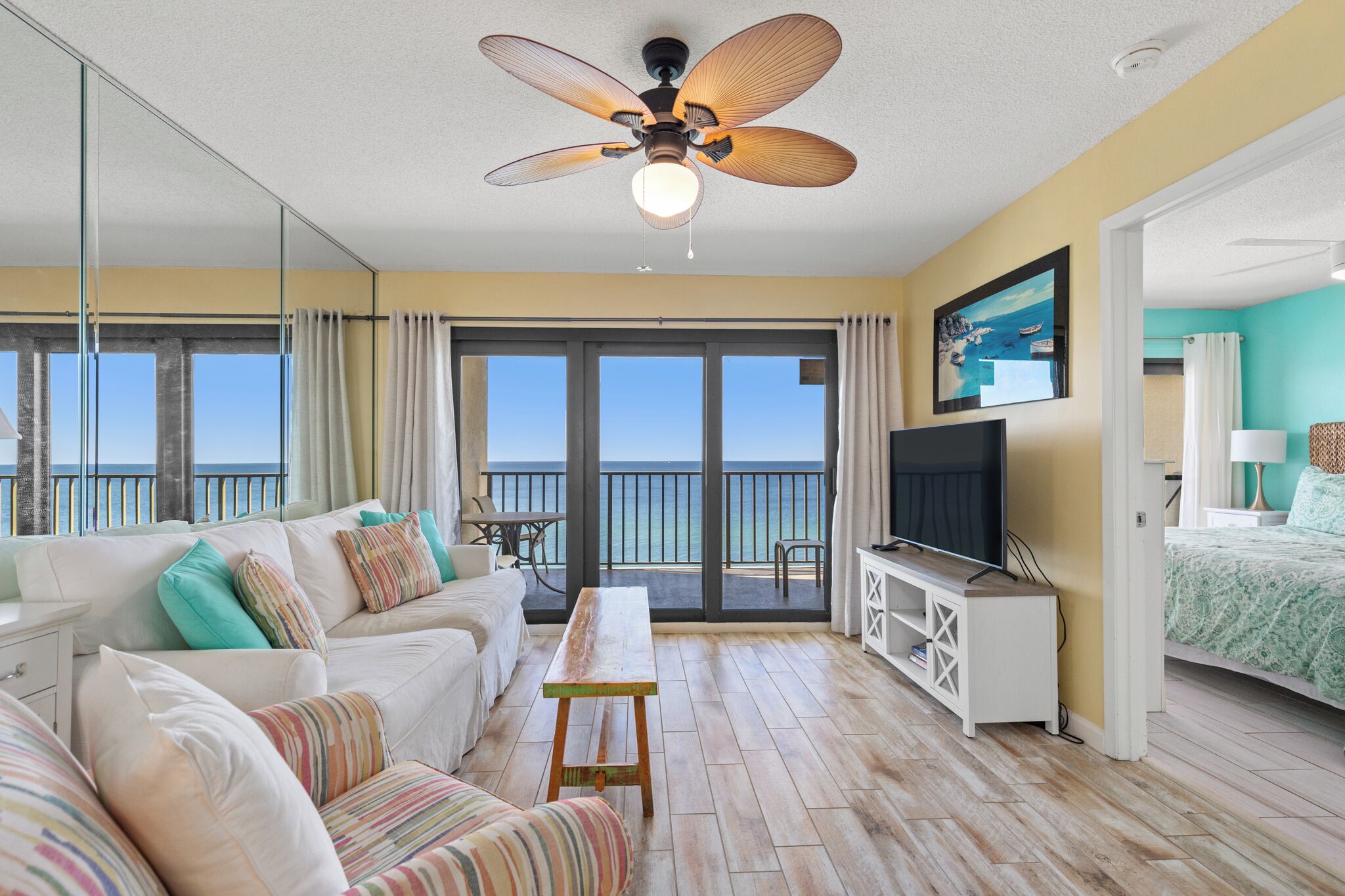 A spacious living room with a large TV, a ceiling fan, and a balcony with a view of the ocean.