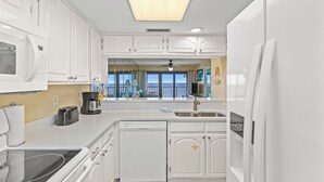 A modern kitchen with white cabinetry, a microwave, toaster, and dishwasher. A large window offers a view of the ocean.