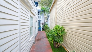 A narrow brick pathway flanked by two buildings with white siding and a small green lawn area with a potted plant.