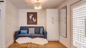 A modern living room with a dark blue sofa, two matching ottomans, a framed artwork above the sofa, white cabinets, and a large window with blinds.