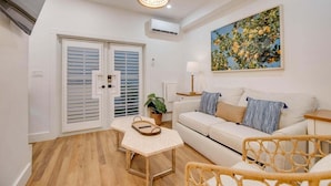 A modern living room with a white sofa, a wooden coffee table, and a wicker chair. There is a painting on the wall, a lamp on a side table, and a window with blinds.