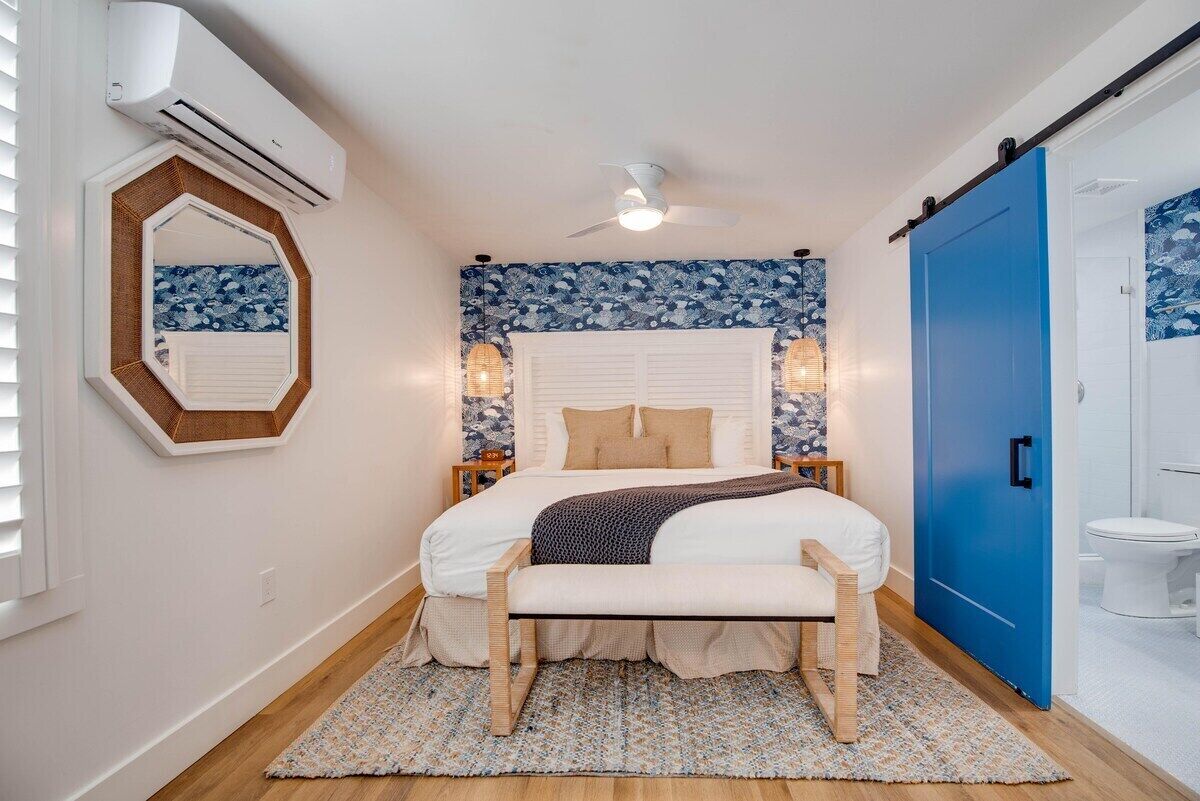 A bedroom with a bed, a bench, a ceiling fan, an air conditioning unit, a mirror, and a blue door.