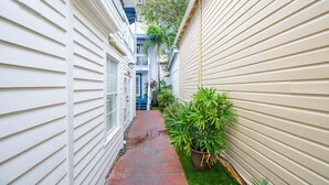 Um caminho de tijolos estreito, ladeado por dois edifícios com revestimento branco e uma pequena área verde com um vaso de planta.