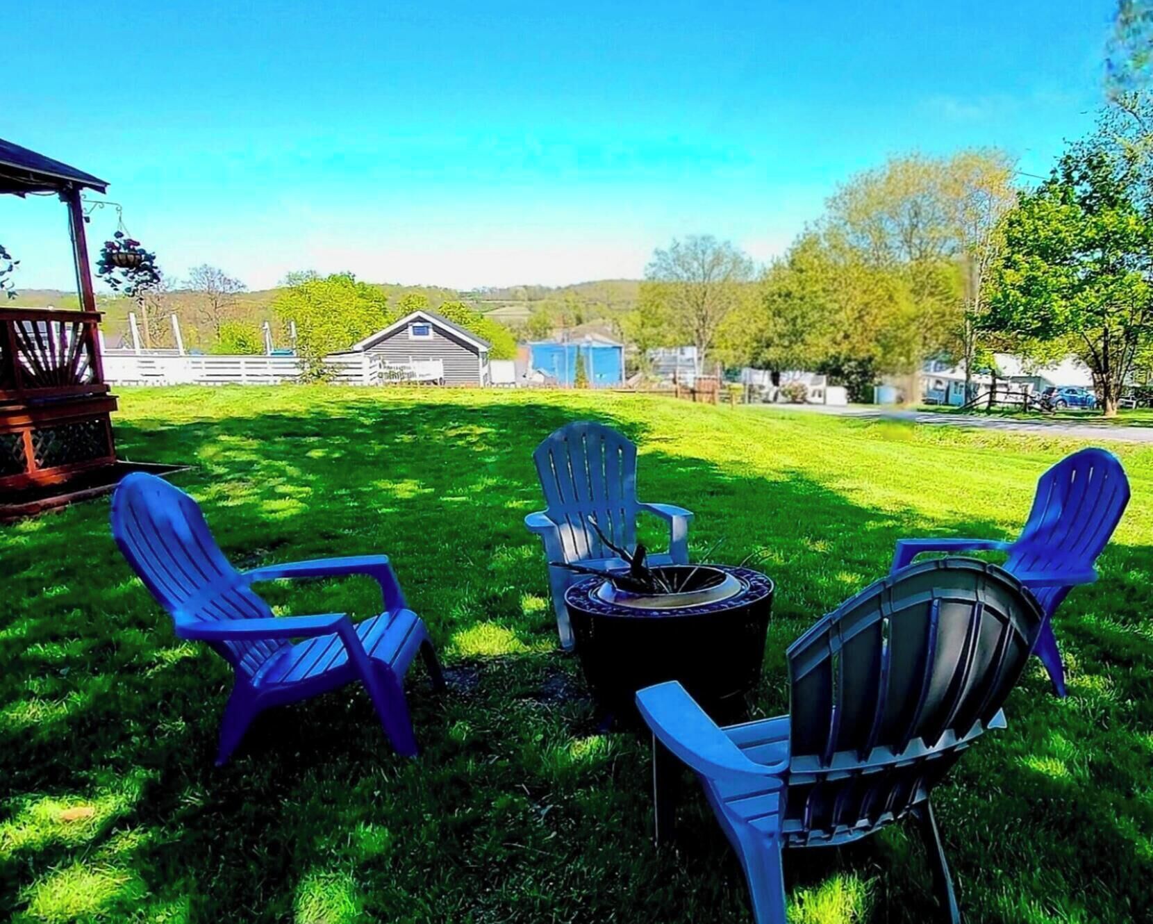 A grassy outdoor area with four blue Adirondack chairs, a metal fire pit, and a wooden pergola.
