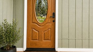 Una puerta principal de madera con un panel de vidrio, un felpudo con el texto 'WELCOME' y una planta en maceta a un lado.