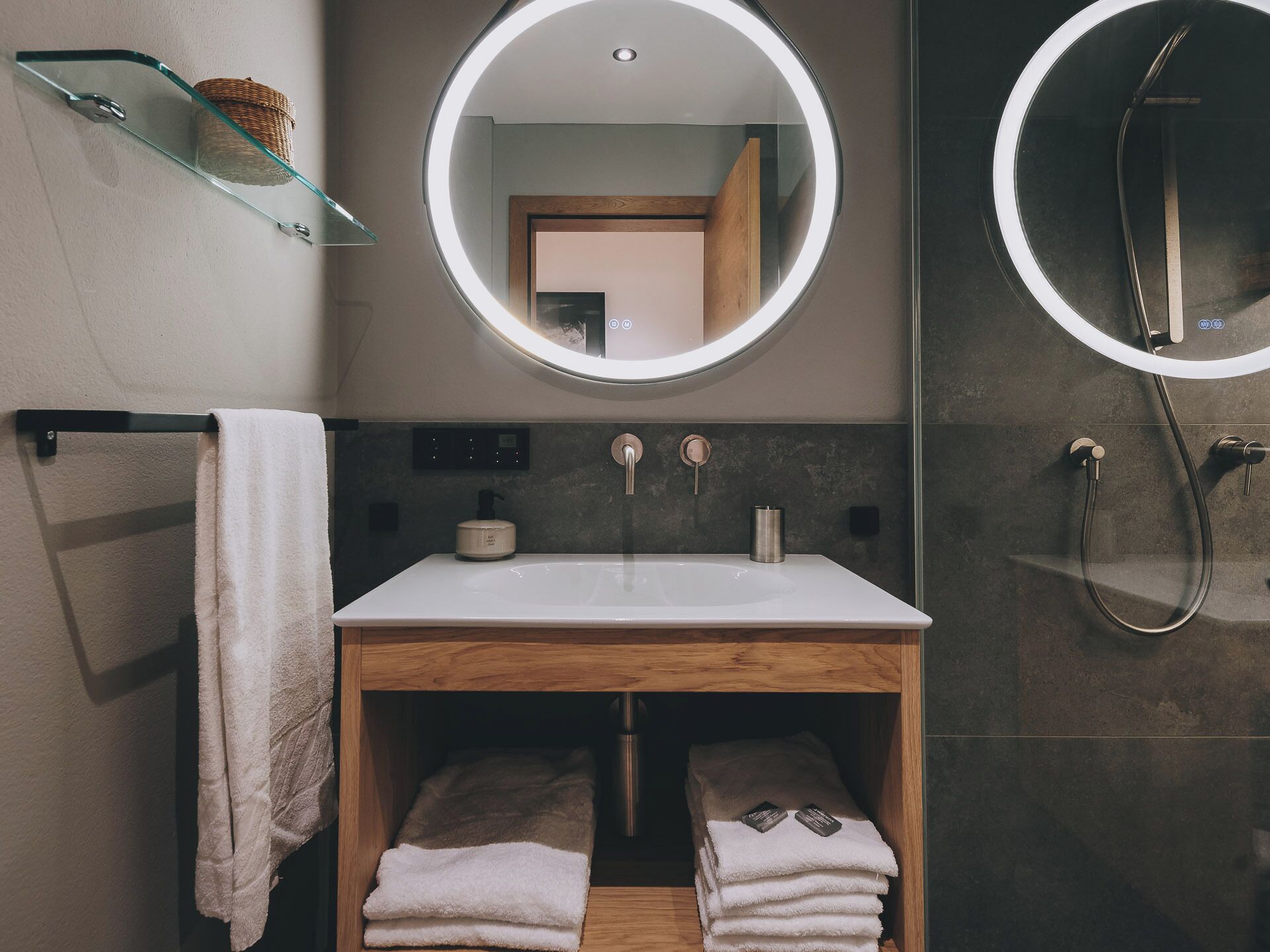 A modern bathroom with a glass shelf, a round mirror, a sink, and a shower.