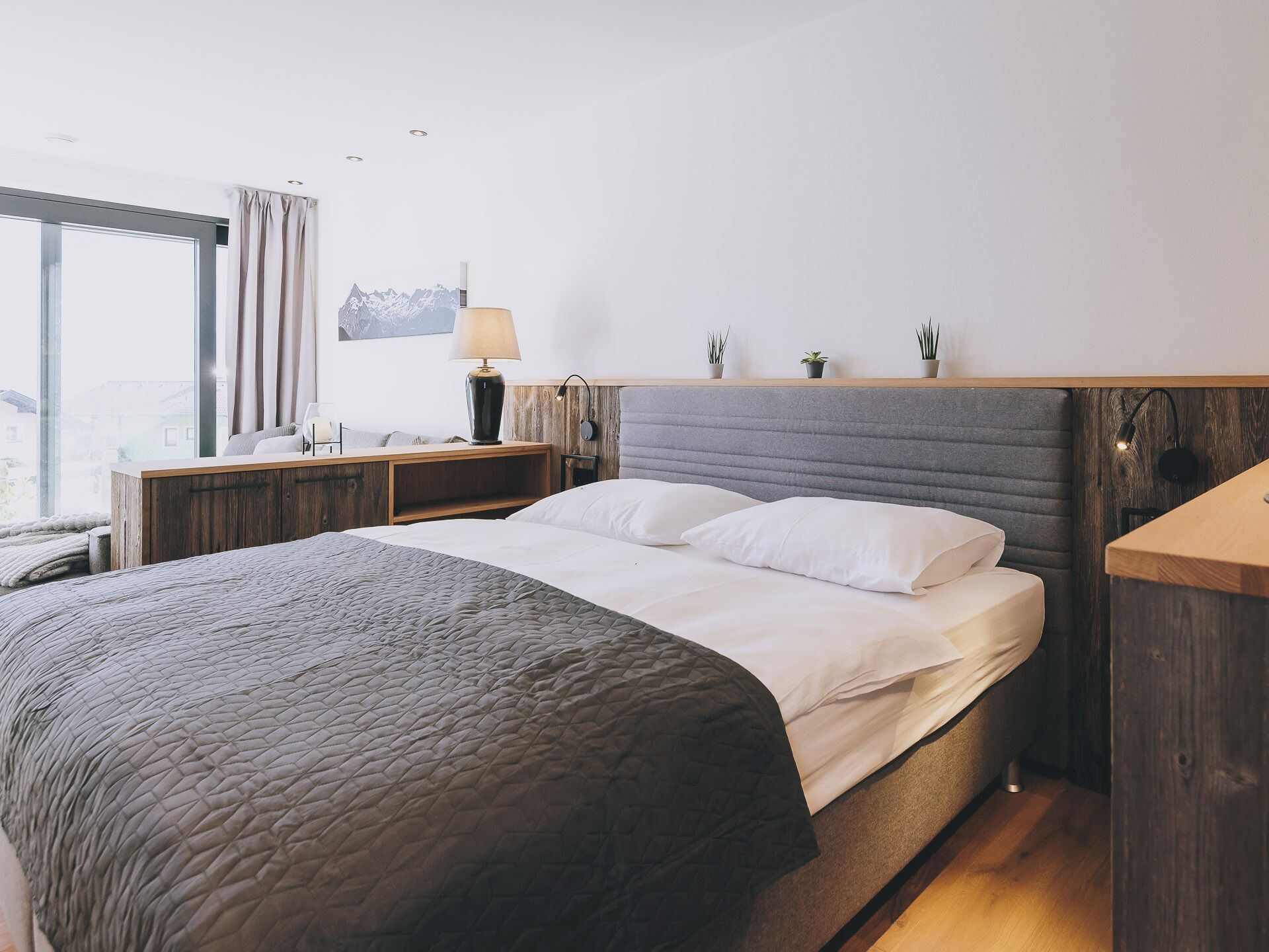 Una habitación de hotel moderna con una cama grande, una mesita de noche de madera, una lámpara y vistas a la ciudad a través de amplios ventanales.