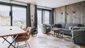 A modern living room with a wooden wall, a grey sofa, a leather chair, and a dining table. - Apartment Alpinero 405 by we rent (Piesendorf)