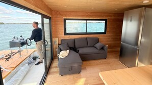 A cabin with a wooden interior, a gray sofa, a refrigerator, and a view of the water through large windows. - Panoramic electric boat, sail on the Lac du Der without a license. Nemours (Éclaron-Braucourt-Sainte-Livière)