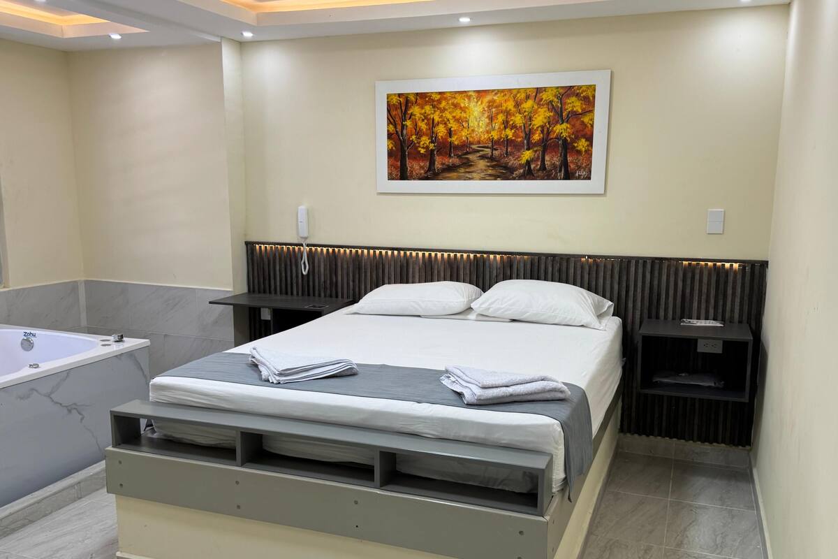 A modern bedroom with a bed, bedside tables, a painting, and a bathroom with a bathtub.