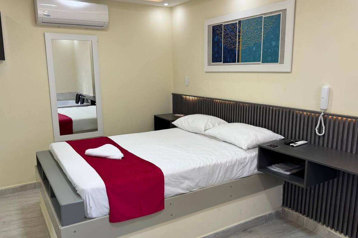 A modern hotel room with a bed, bedside table, and a wall-mounted air conditioner.