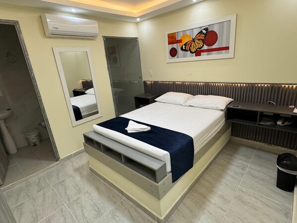 A modern hotel room with a large bed, a bedside table, a mirror, an air conditioning unit, and a bathroom with a toilet and sink. - HOTEL MAGISTRAL SUITER (Quibdó)