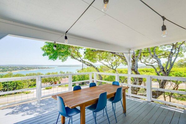 Un área de comedor al aire libre cubierta, con una mesa de madera y sillas azules, con vista a un paisaje de árboles y una masa de agua.
