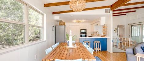 A spacious dining area with a wooden table and chairs, a hanging light fixture, and large windows offering a view of the outdoors.