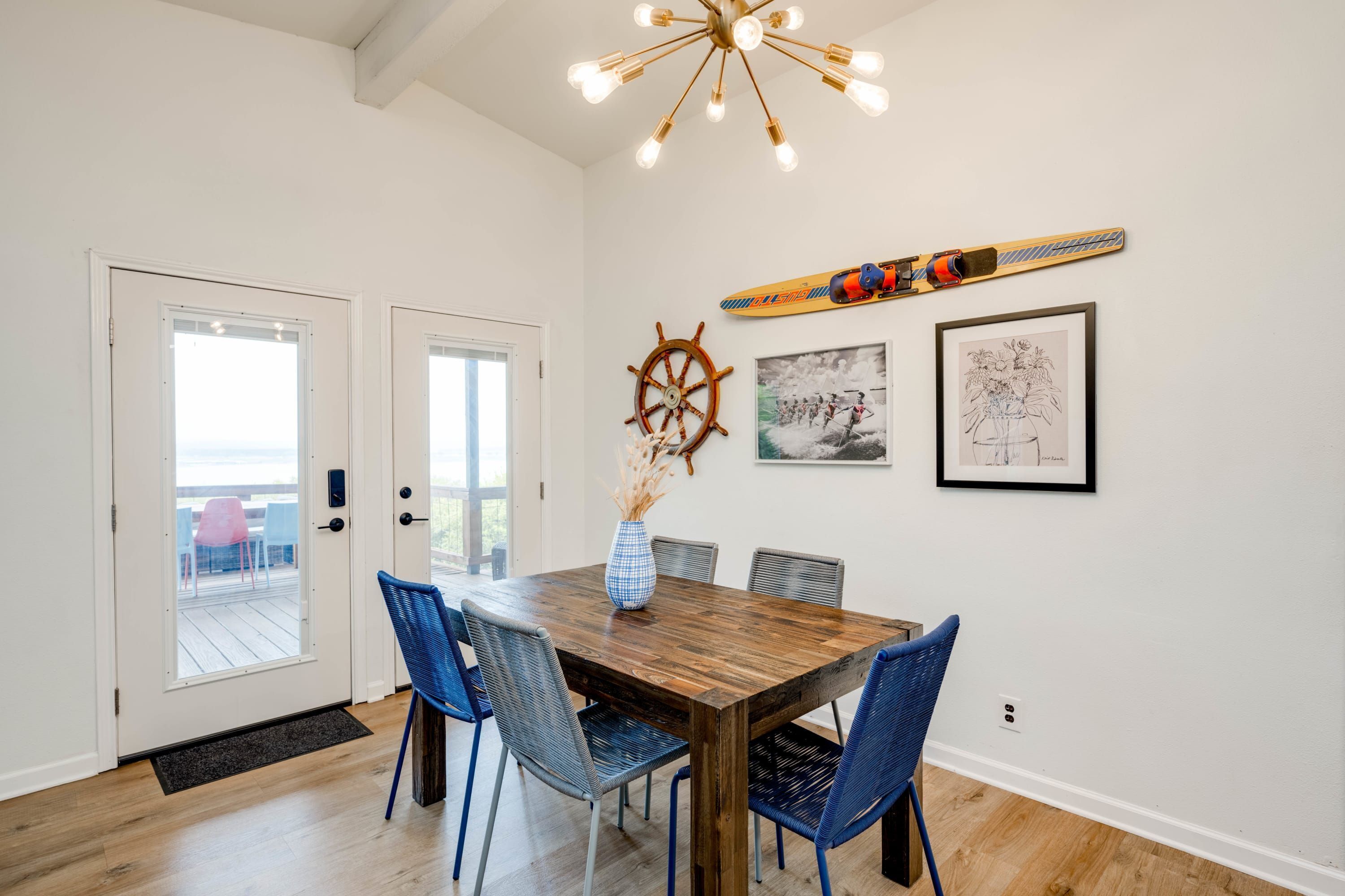 A dining area with a wooden table, blue chairs, a ship's wheel, and framed artwork on the wall.