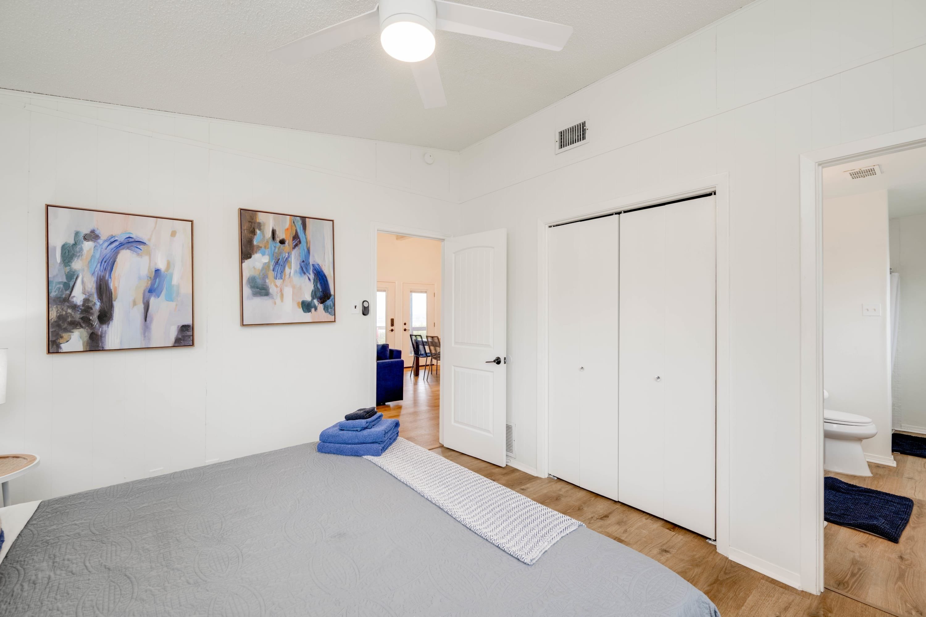 A bedroom with a bed, two abstract paintings, a ceiling fan, and a door leading to another room.