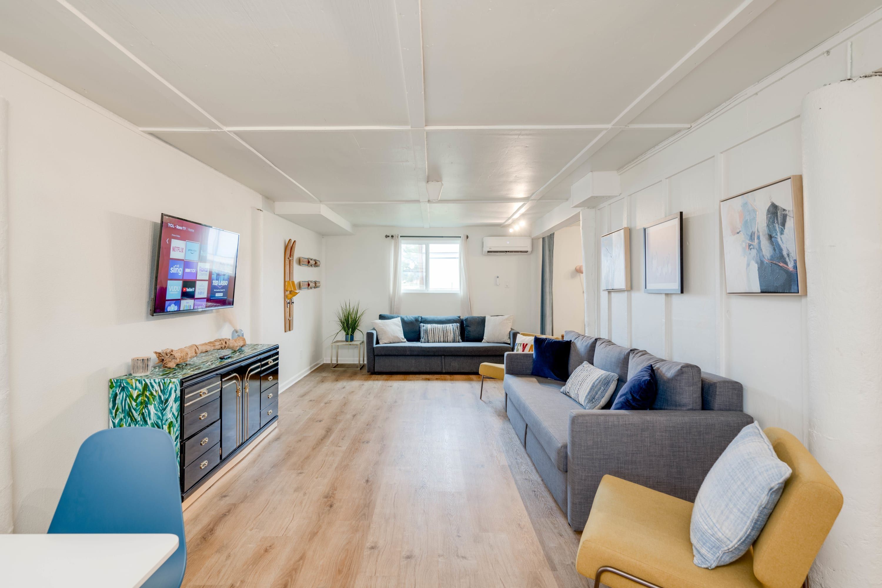 Een moderne woonkamer met een grijze bank, een gele fauteuil en een blauwe stoel. Er is een flatscreen televisie aan de muur, een bijzettafeltje met een plant en een kast met decoratieve objecten.