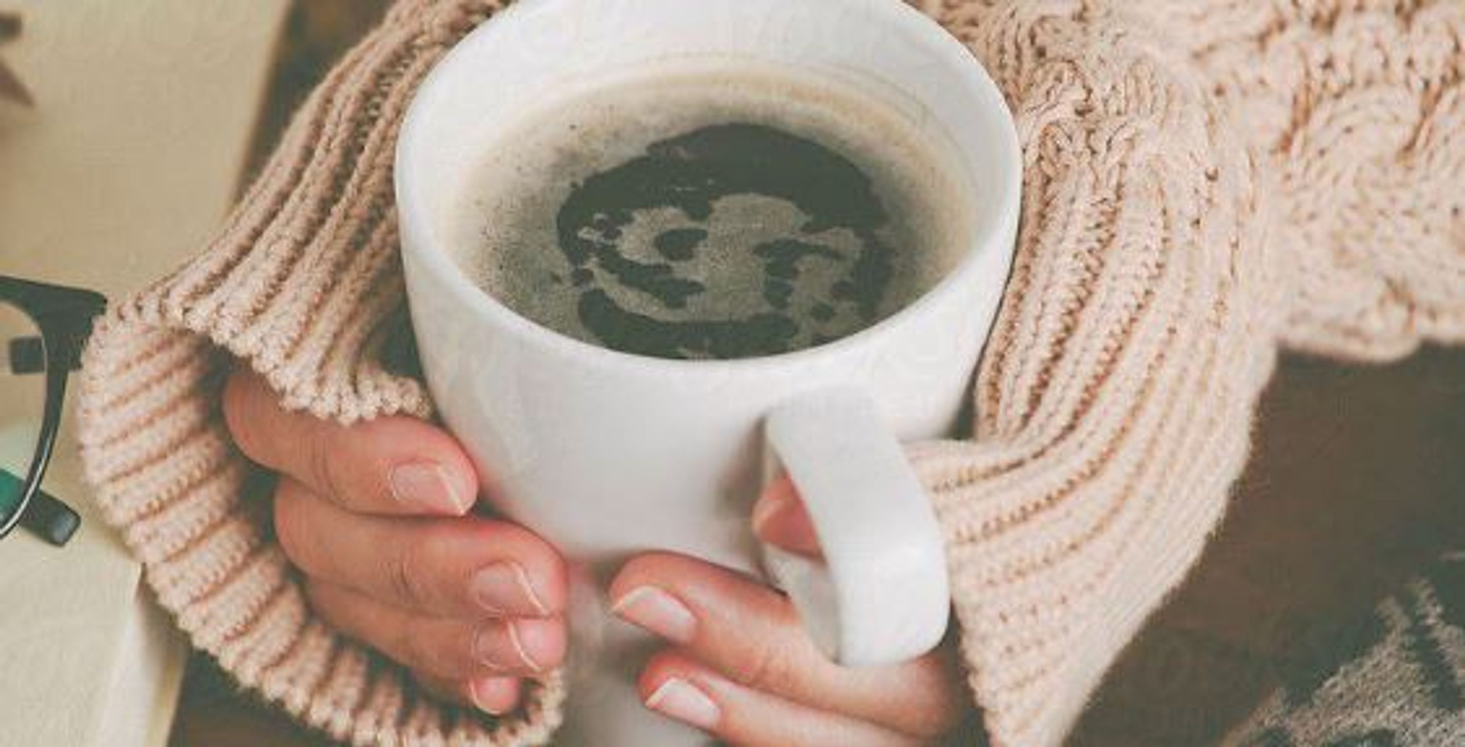 A person holding a white mug with a dark liquid, possibly coffee or tea, and wearing a knitted garment.