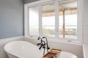 A bathroom with a freestanding bathtub, a window with a view of the outdoors, and a wooden tray on the floor.
