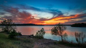 A sunset over a calm lake with trees in the foreground and hills in the distance.