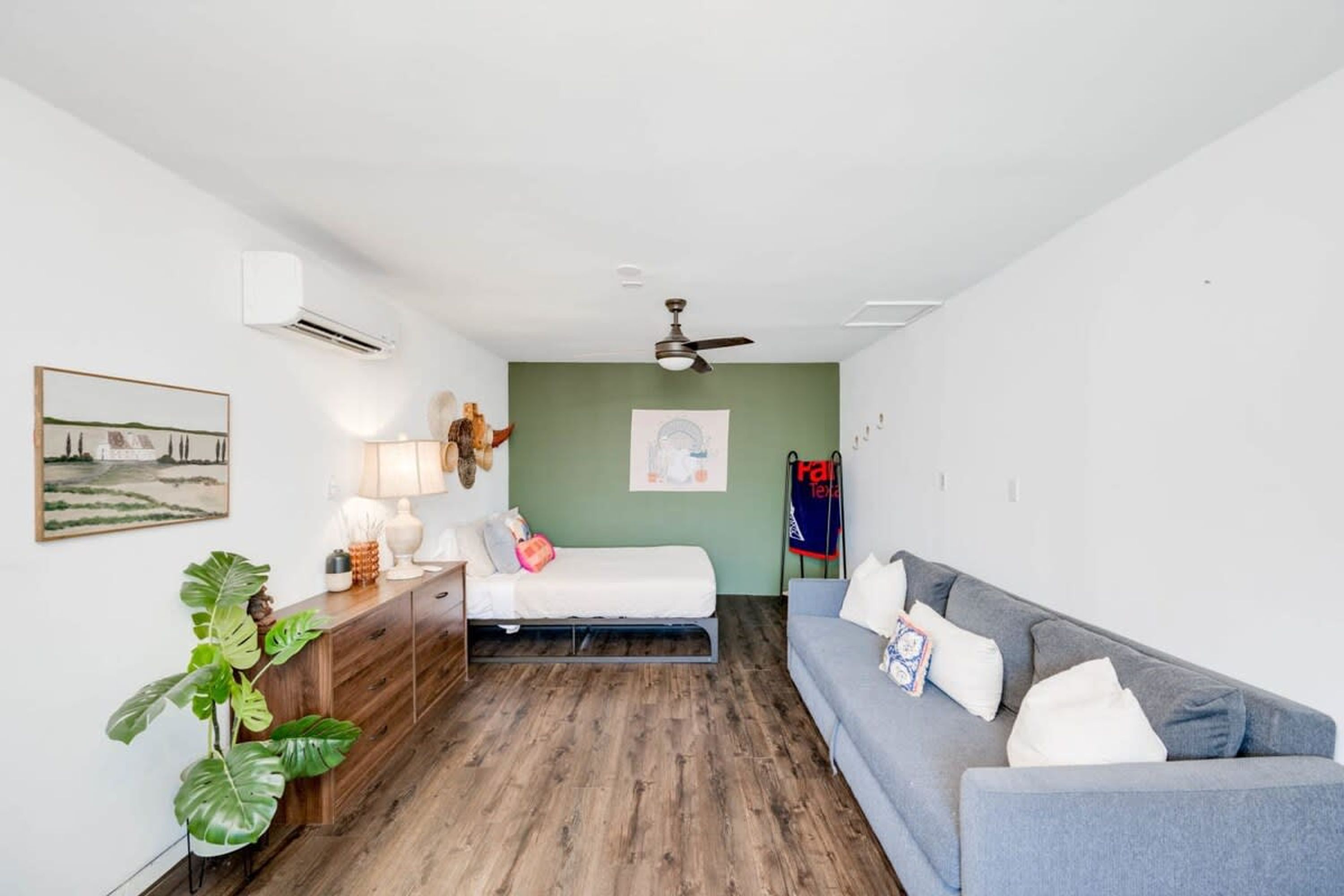 Een kamer met een bed, een bank, een bureau, een lamp, een plant en een plafondventilator.