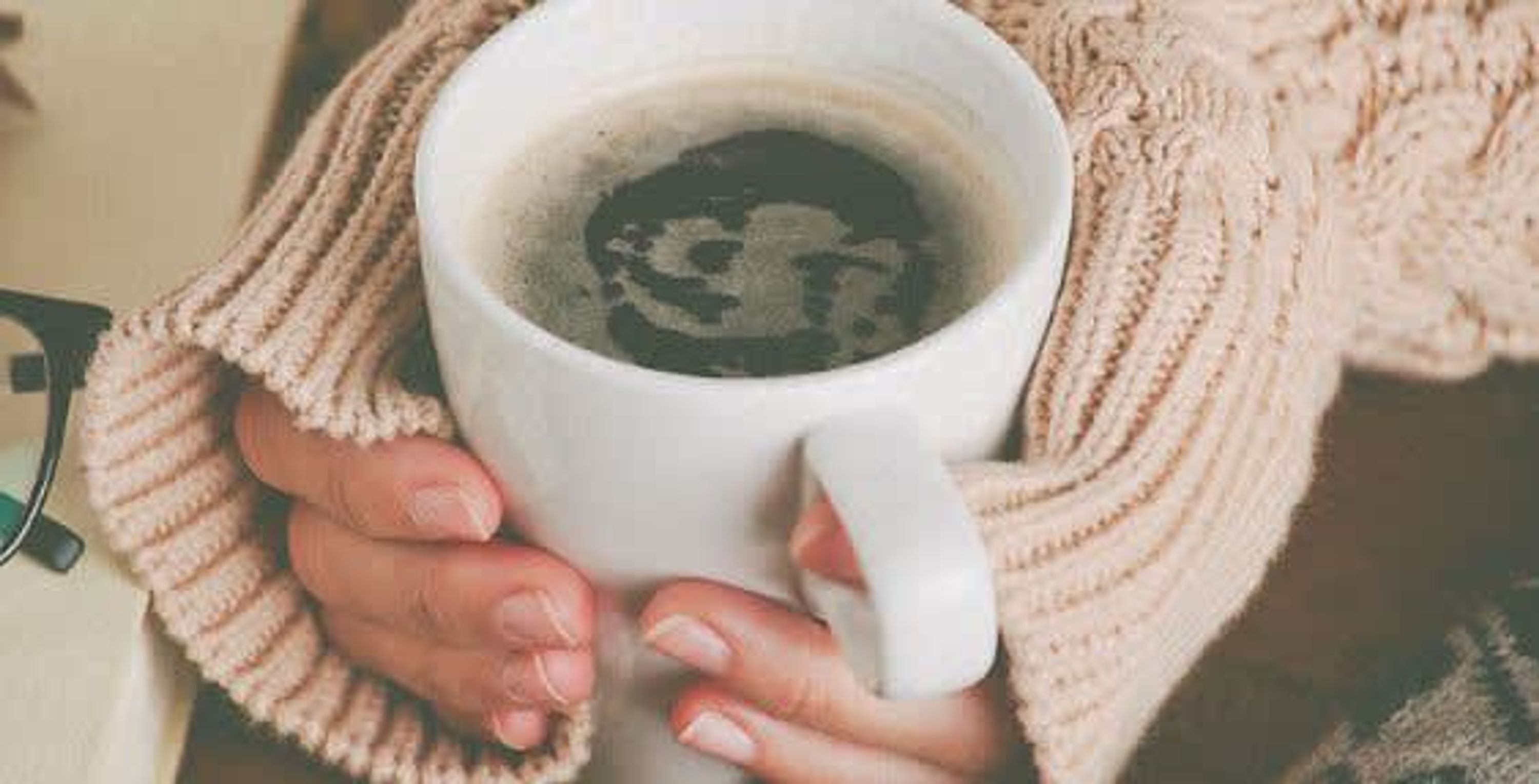 En person holder en hvid kop med en mørk væske, muligvis kaffe eller te, iført en beige strikket trøje.