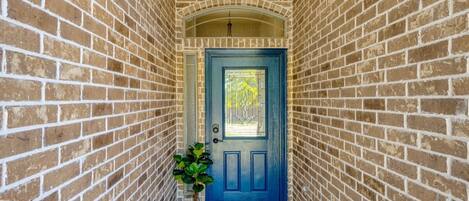 A narrow brick corridor leading to a blue door with a glass panel, a potted plant, and a 'Hello' mat.