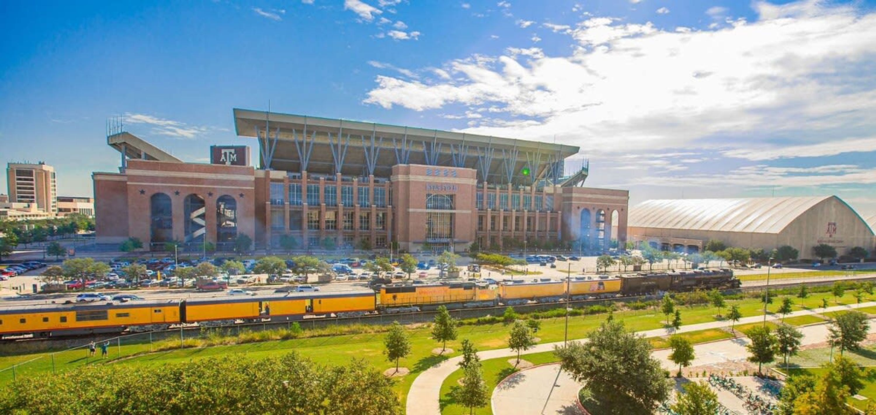 A large building with a flat roof and multiple windows, surrounded by a parking lot and a train track.