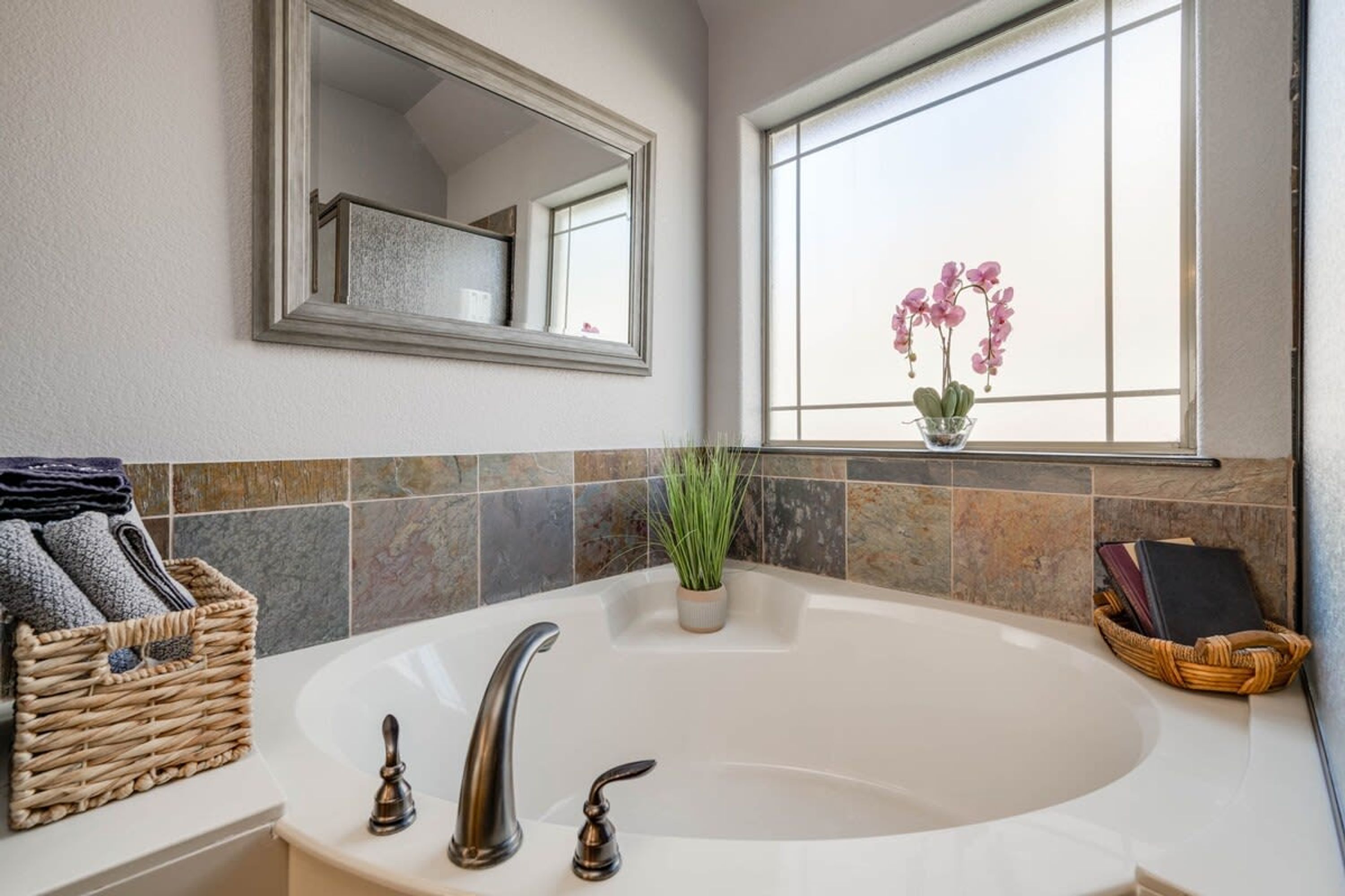 Une salle de bain avec une grande baignoire, un miroir, une fenêtre agrémentée d’une plante orchidée et un panier en osier contenant des serviettes.