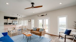 Amplia sala de estar con un tapete azul, un sofá marrón y un diván azul. Se observa un ventilador de techo, una mesa de comedor con sillas y una zona de cocina al fondo.