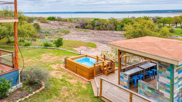 Un jacuzzi con cubierta de madera, un área de estar techada con sillas azules y vista a un lago.