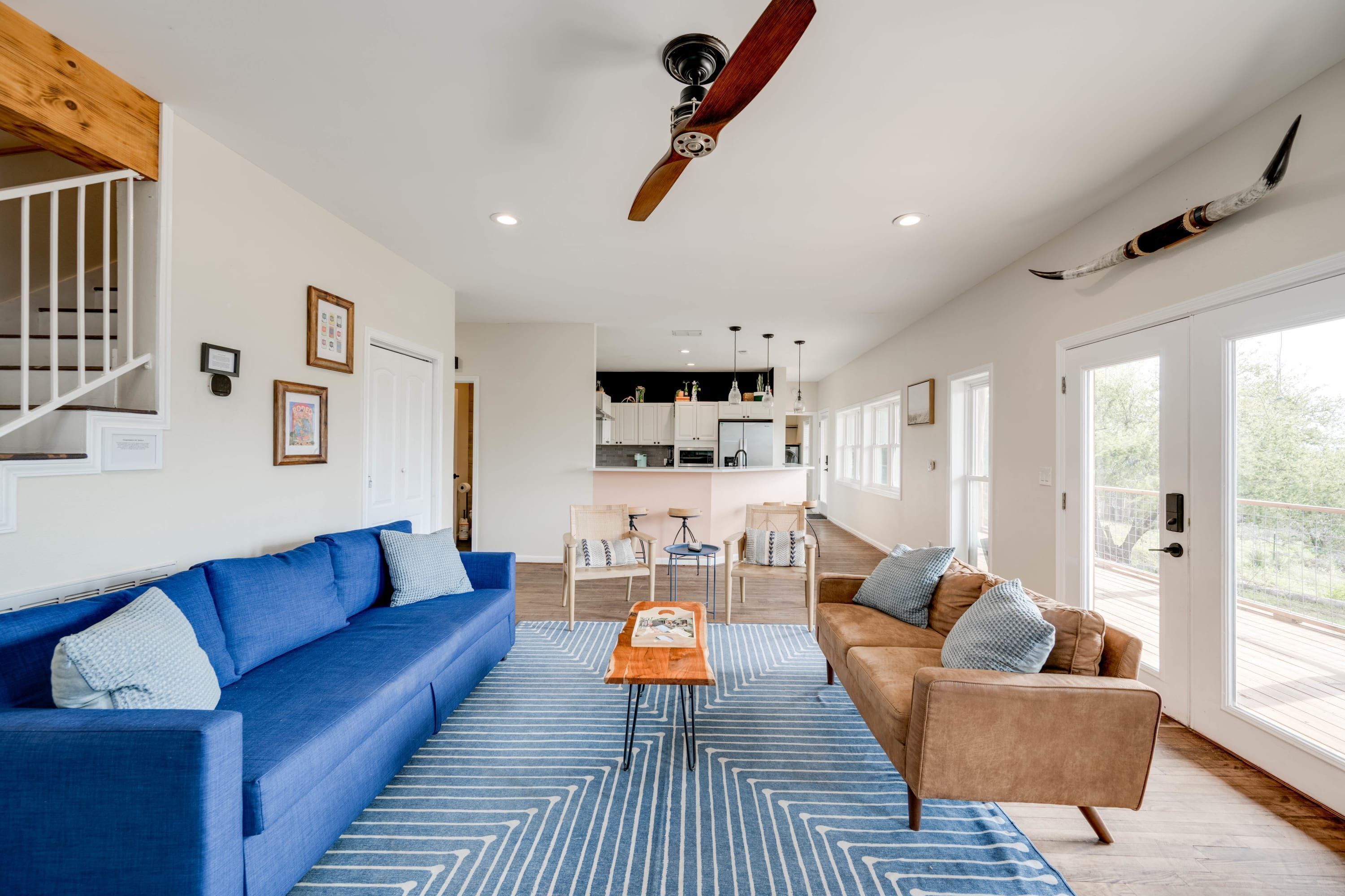 A spacious living area with a blue sofa, a brown sofa, and a wooden coffee table. There is a staircase with a wooden railing on the left and a large window on the right.
