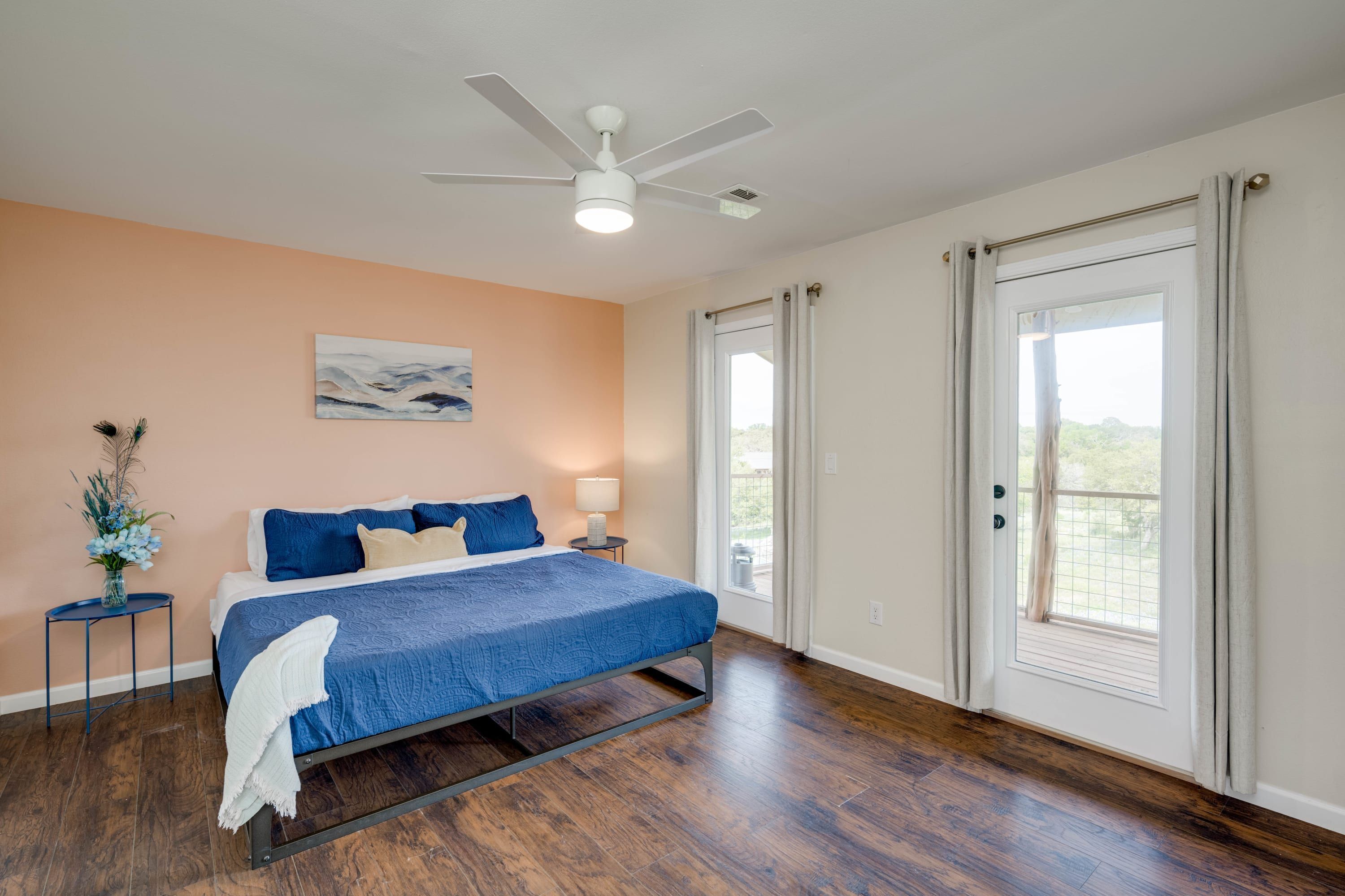 Une chambre à coucher avec un lit, un ventilateur de plafond, une fenêtre avec des rideaux, une table de chevet avec une lampe et un tableau au mur.