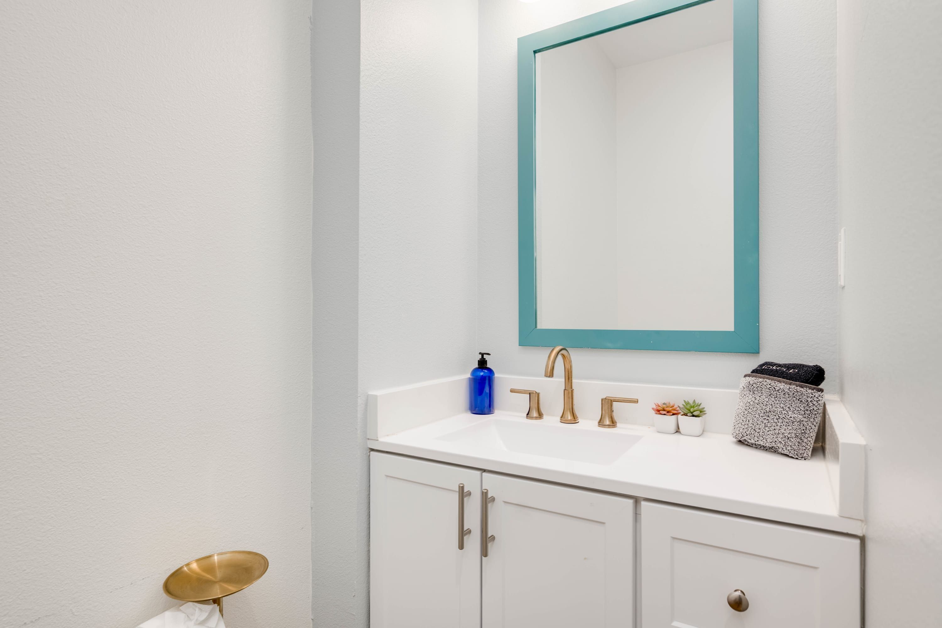 A bathroom with a white sink, gold faucets, a blue soap dispenser, and a small potted plant.
