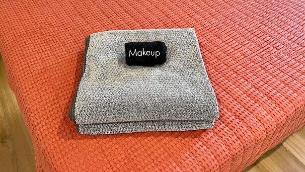 A neatly folded grey blanket with a black tag labeled "Makeup" on a red textured ottoman.