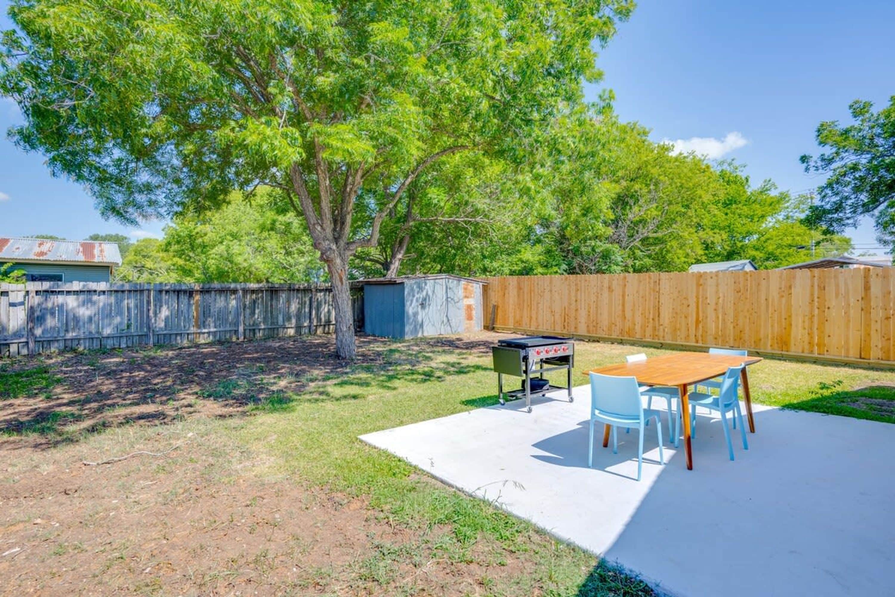 Un patio trasero con cerco de madera, un árbol, una parrilla y una mesa con sillas.