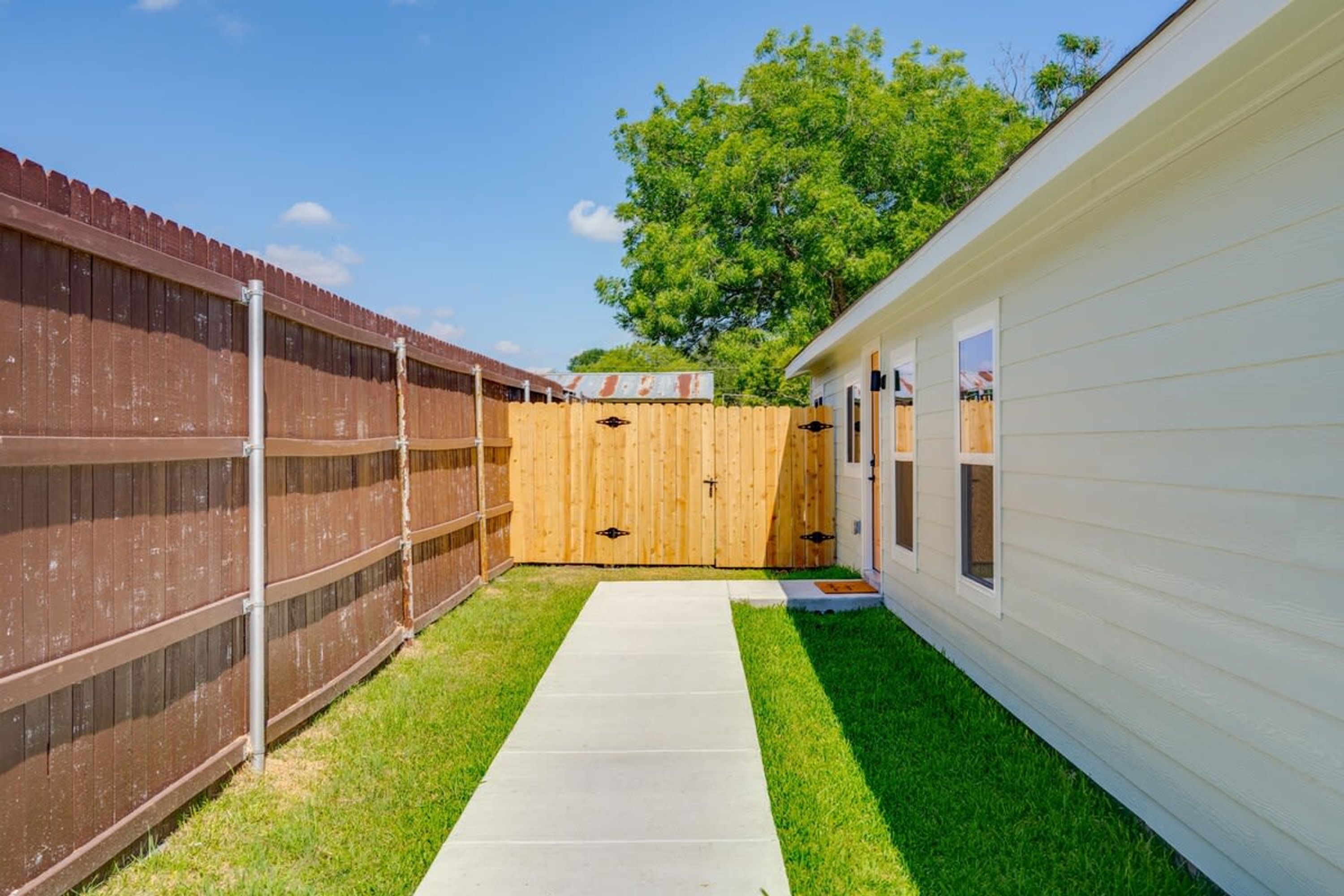 A fenced backyard with a concrete pathway leading to a wooden gate, a white building with windows, and a tree in the background.