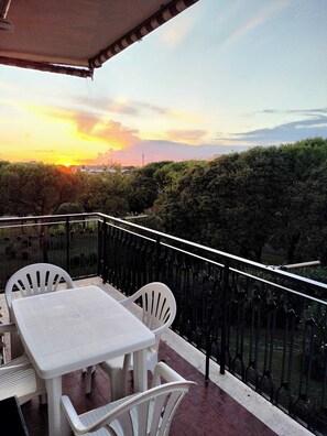 Ein Balkon mit Tisch und Stühlen, mit Blick auf einen Sonnenuntergang, im Hintergrund Bäume und Gebäude.