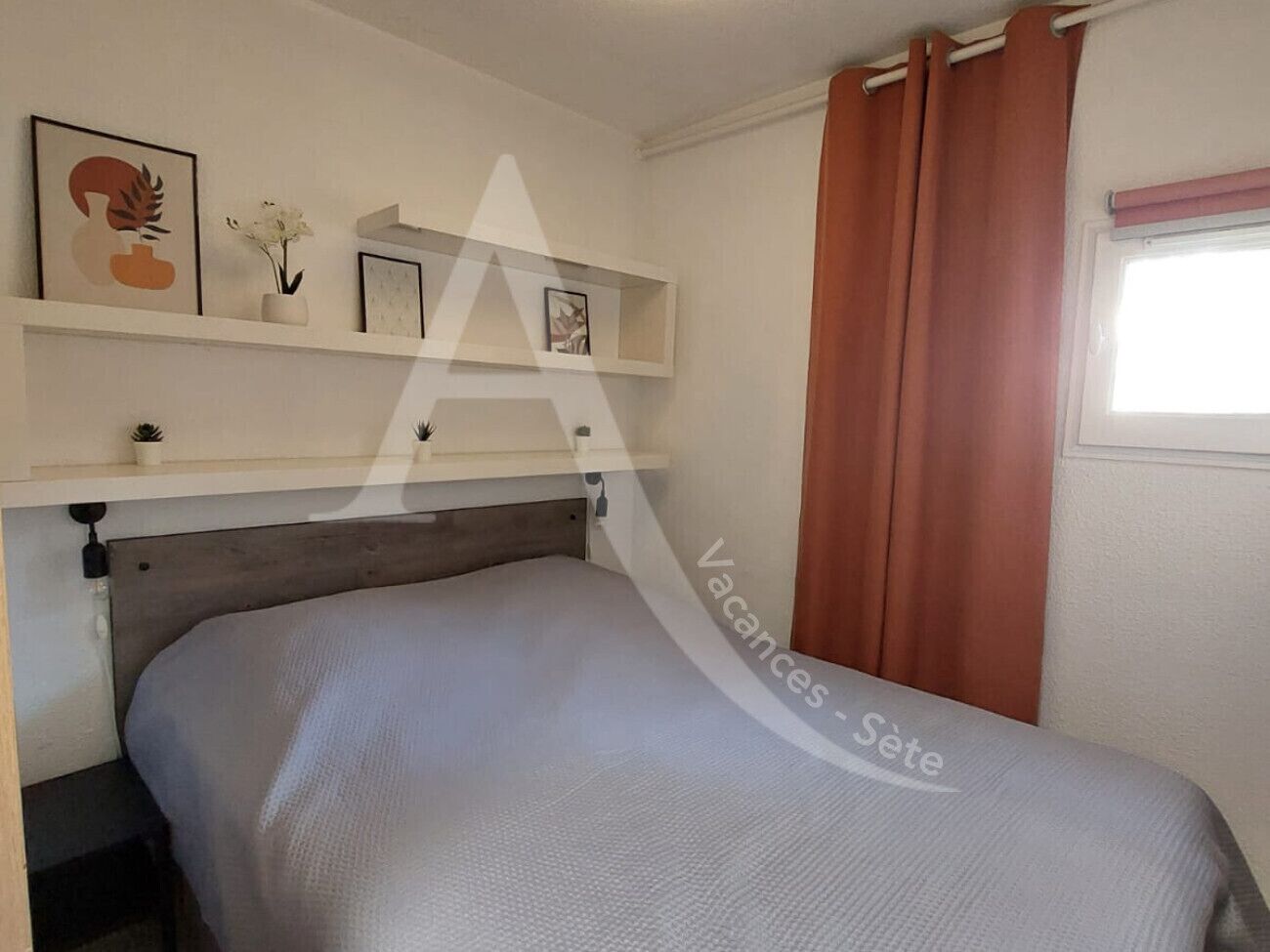 A bedroom with a bed, a shelf with framed pictures, a window with curtains, and a wall-mounted light fixture.