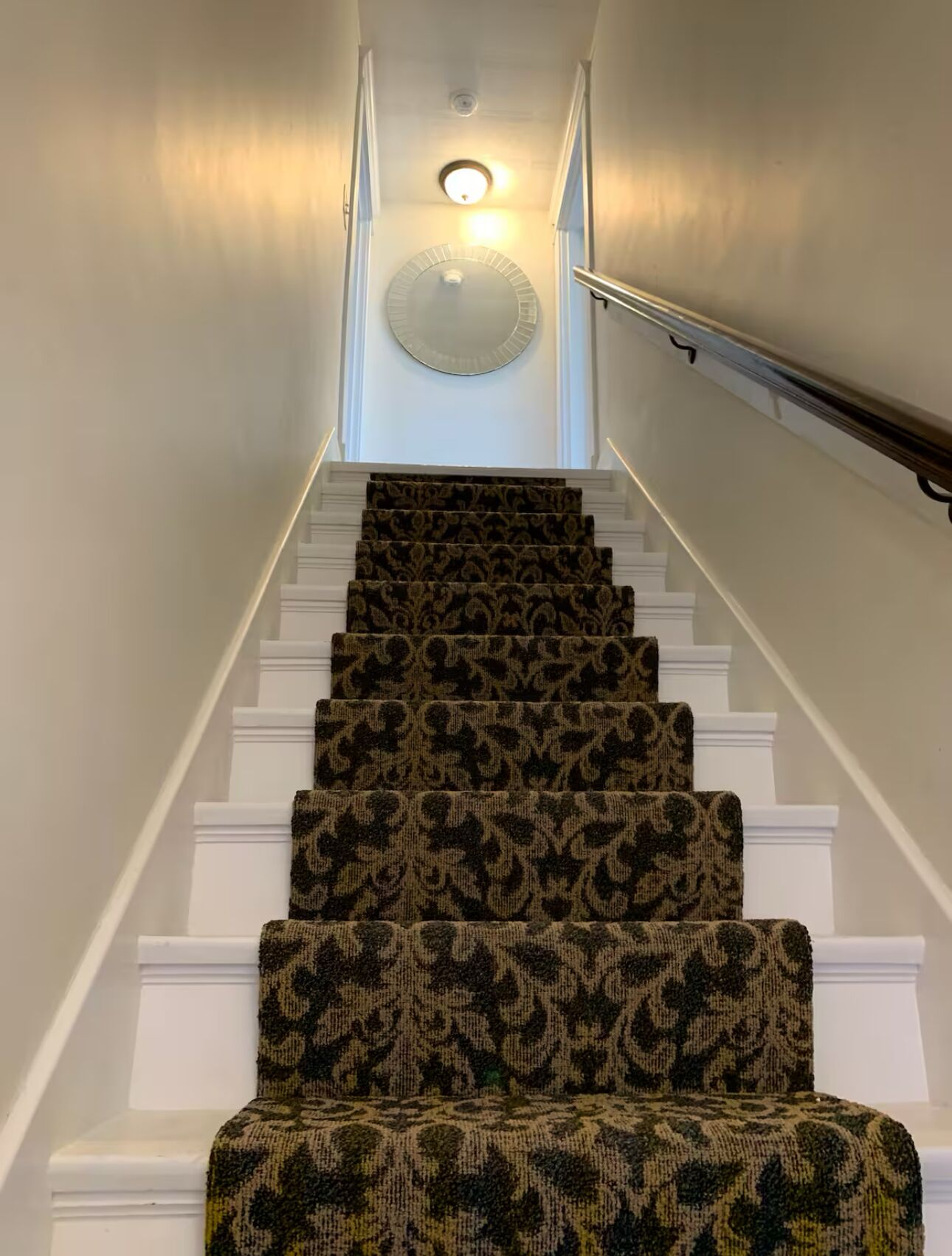 A staircase with a patterned carpet, white balusters, and a wall-mounted light fixture.