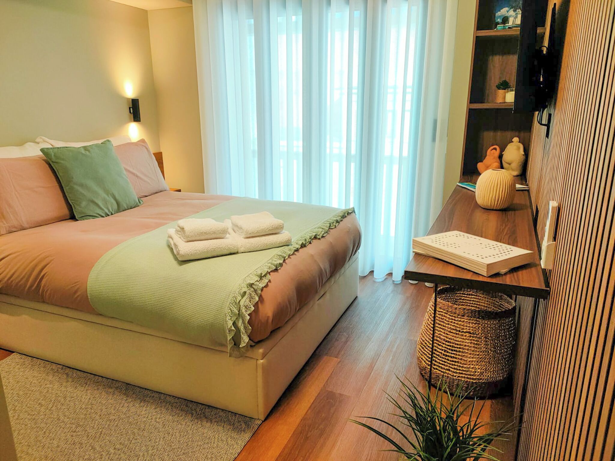 Un dormitorio ordenado con cama, mesita de noche, lámpara y una pequeña estantería con objetos decorativos.