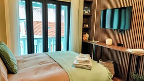 Una camera d'hotel con un letto, una TV fissata al muro, una finestra con vista sugli edifici e uno scaffale con vari oggetti.