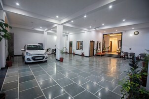 Um hall amplo com um carro branco estacionado no centro, plantas em vasos e uma escadaria que leva a outra área.