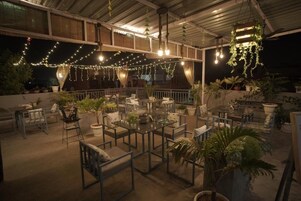 Un espacio al aire libre con mesas y sillas, luces colgantes y plantas en macetas.