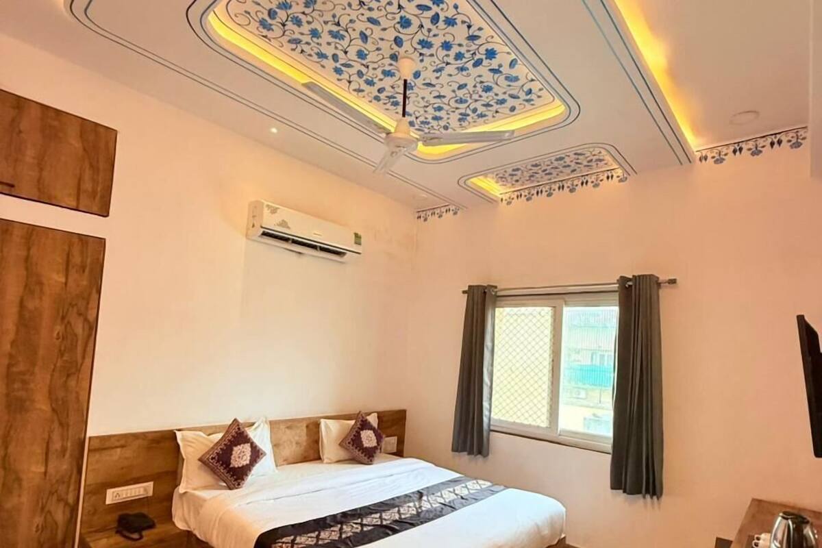 A bedroom with a wooden bed, a ceiling with floral patterns, a window with curtains, and a small table with a kettle.