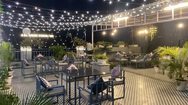 An outdoor seating area with tables and chairs, string lights overhead, and potted plants.