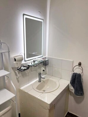 Un baño con un lavamanos blanco, un espejo con luz, un perchero y un secador de pelo en un estante.