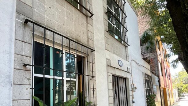 Edifici de dues plantes amb grans finestres i una porta, envoltat d'arbres i una vorada.
